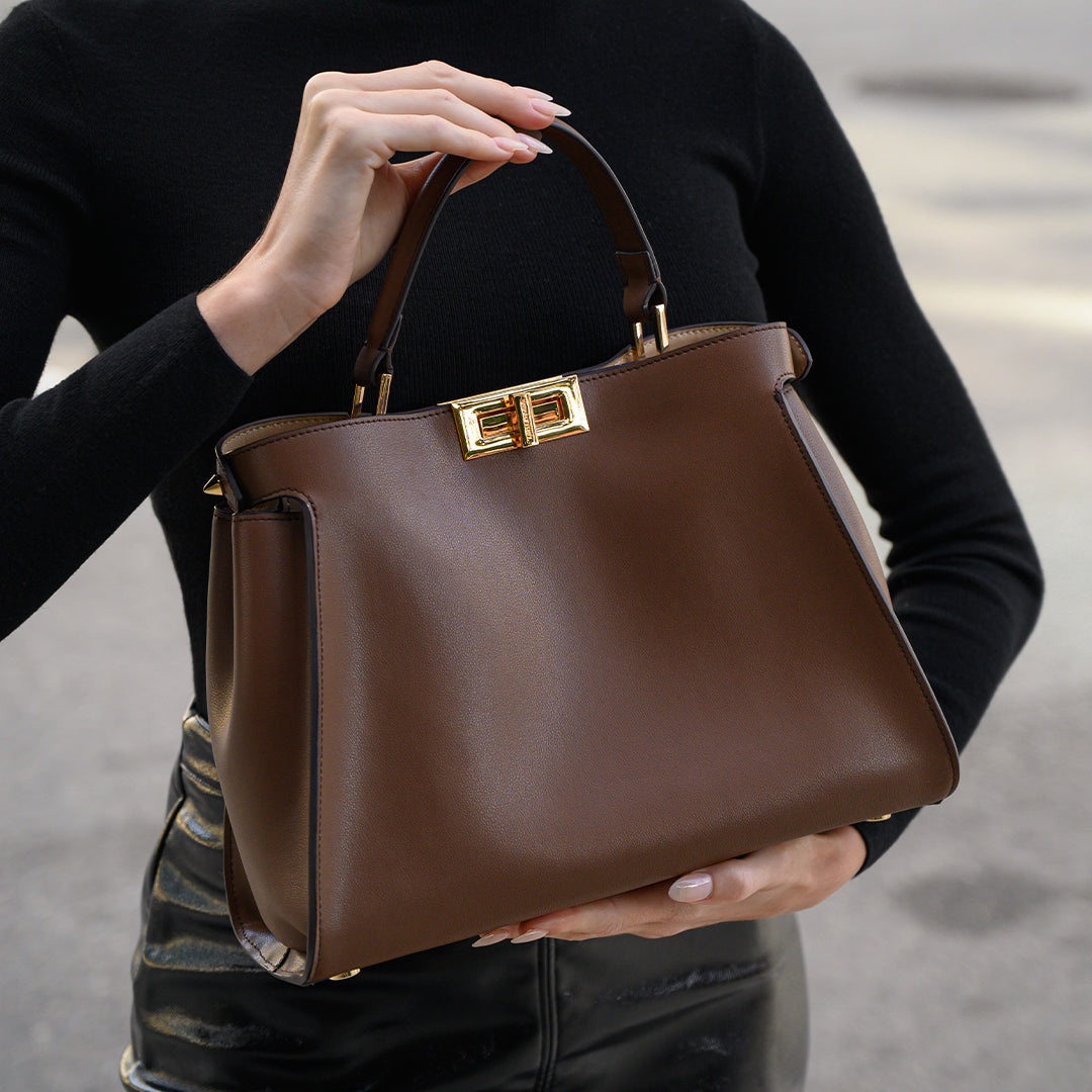 Brown leather handbag with gold clasp held by a person wearing a black outfit.